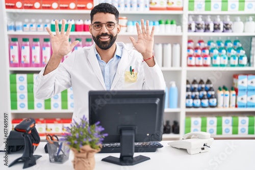 Hispanic man with beard working at pharmacy drugstore showing and pointing up with fingers number ten while smiling confident and happy.