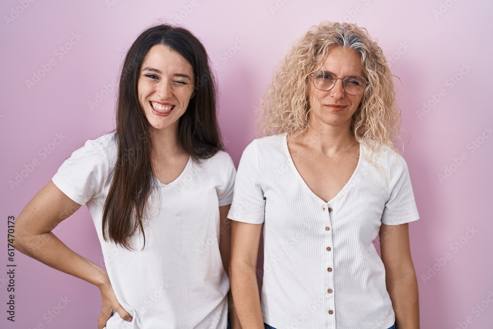 Mother and daughter standing together over pink background winking looking at the camera with sexy expression, cheerful and happy face. Mother and daughter standing together over pink background winking looking at the camera with sexy expression, cheerful and happy face.