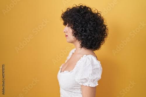 Young brunette woman with curly hair standing over yellow background looking to side, relax profile pose with natural face and confident smile.