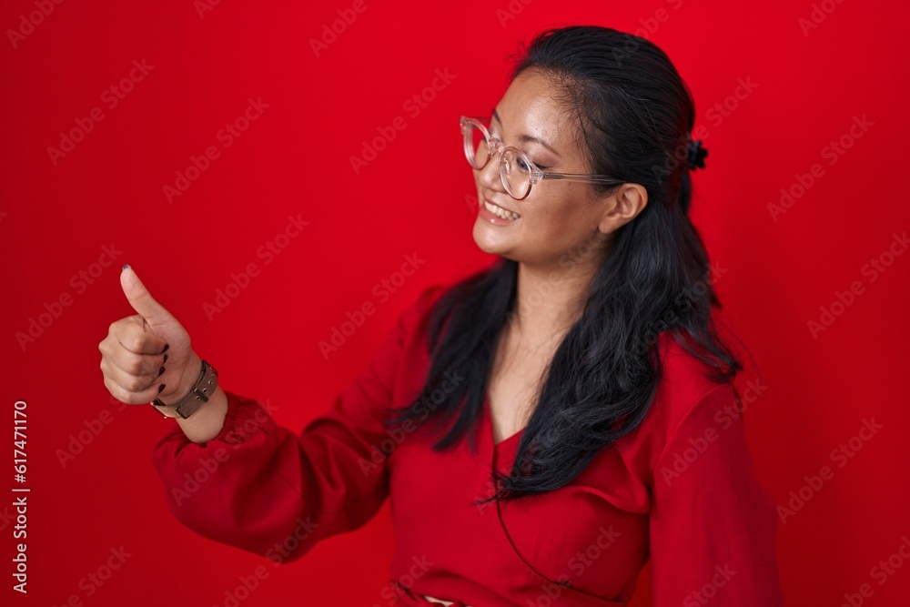 Asian young woman standing over red background looking proud, smiling doing thumbs up gesture to the side Asian young woman standing over red background looking proud, smiling doing thumbs up gesture to the side