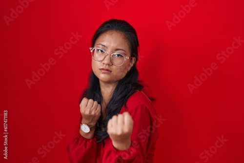 Asian young woman standing over red background ready to fight with fist defense gesture, angry and upset face, afraid of problem