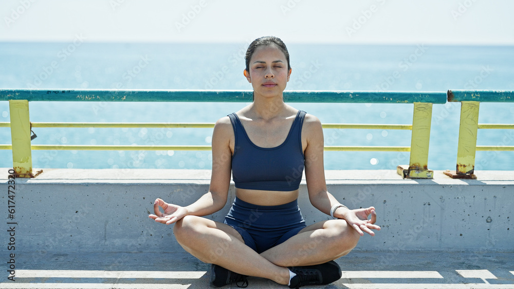 Young beautiful hispanic woman doing yoga exercise sitting on bench at seaside