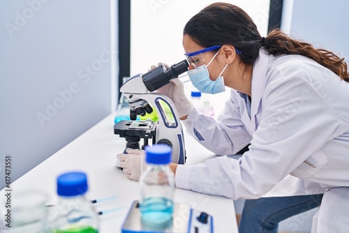 Young hispanic woman wearing scientist uniform using microscope at laboratory