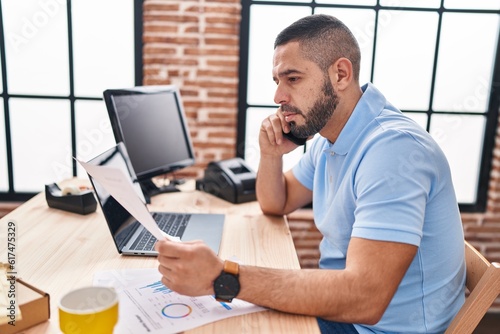 Young latin man ecommerce business worker reading document talking on smartphone at office