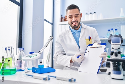Hispanic man working at scientist laboratory holding blank clipboard smiling with a happy and cool smile on face. showing teeth.