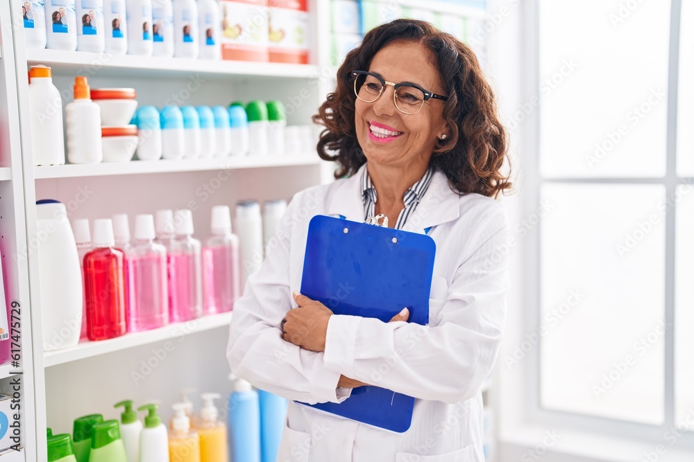 Middle age woman pharmacist smiling confident holding clipboard at pharmacy