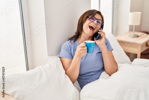 Middle age woman talking on smartphone drinking coffee at bedroom