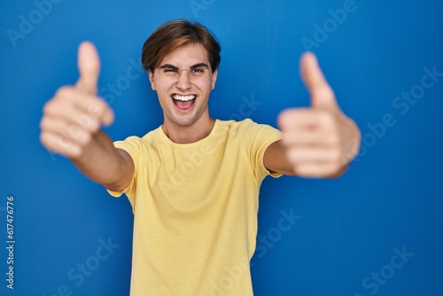 Young man standing over blue background approving doing positive gesture with hand, thumbs up smiling and happy for success. winner gesture. © Krakenimages.com