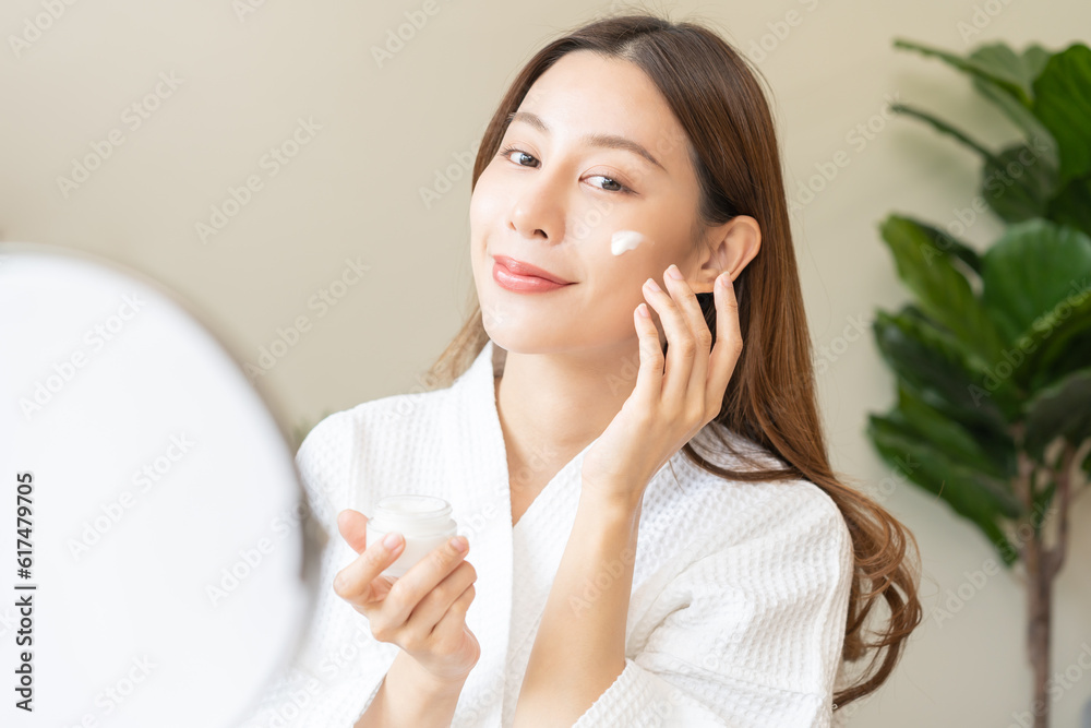 Facial beauty skin care, smile of pretty asian young woman in bathrobe looking at mirror, hand applying moisturizer lotion on her face, holding jar of skin cream before makeup cosmetic routine at home Facial beauty skin care, smile of pretty asian young woman in bathrobe looking at mirror, hand applying moisturizer lotion on her face, holding jar of skin cream before makeup cosmetic routine at home