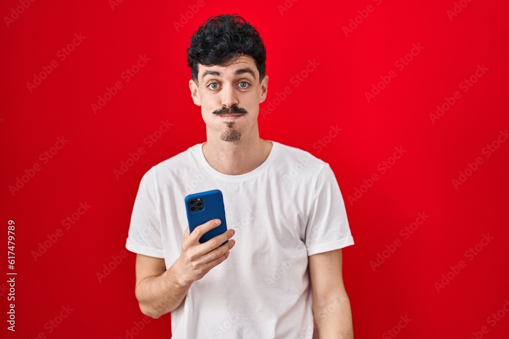 Hispanic man using smartphone over red background puffing cheeks with funny face. mouth inflated with air, crazy expression. Hispanic man using smartphone over red background puffing cheeks with funny face. mouth inflated with air, crazy expression.