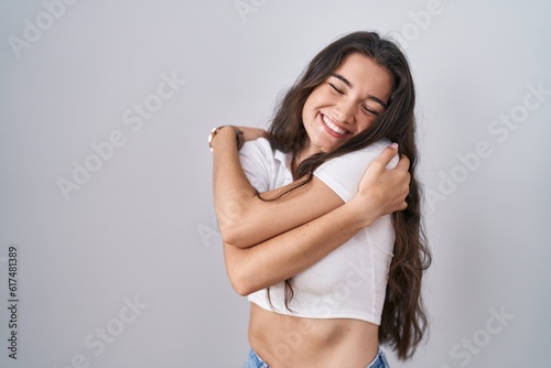 Young teenager girl standing over white background hugging oneself happy and positive, smiling confident. self love and self care