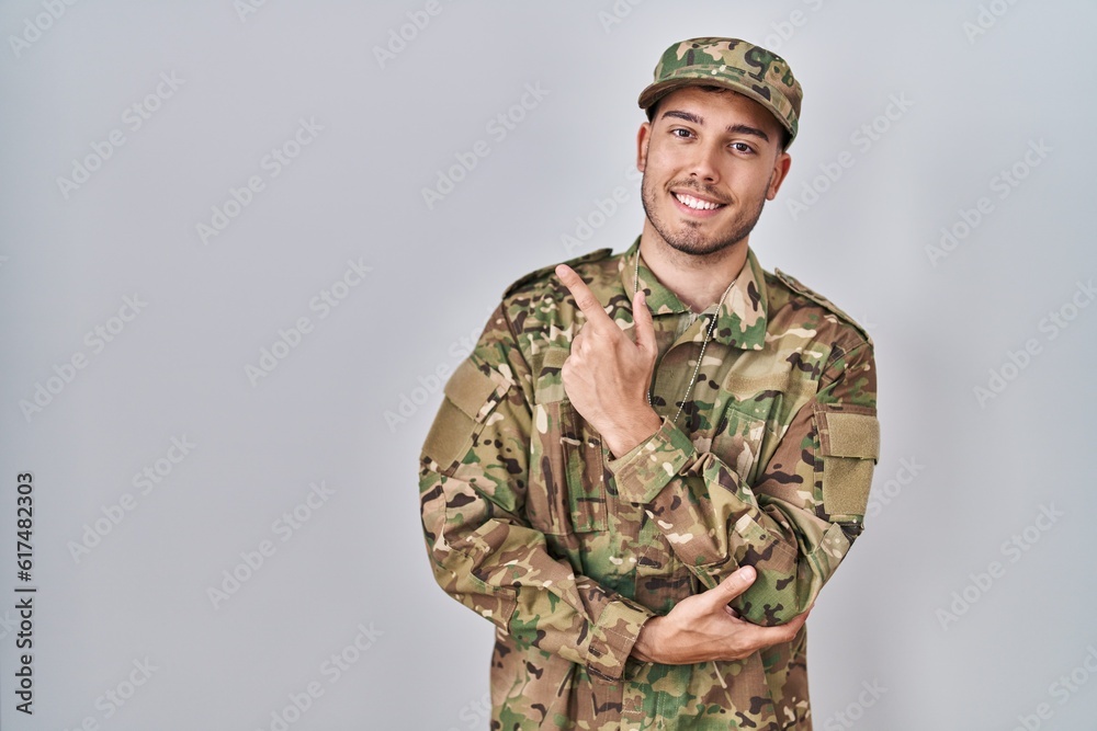 Young hispanic man wearing camouflage army uniform with a big smile on face, pointing with hand and finger to the side looking at the camera. Young hispanic man wearing camouflage army uniform with a big smile on face, pointing with hand and finger to the side looking at the camera.