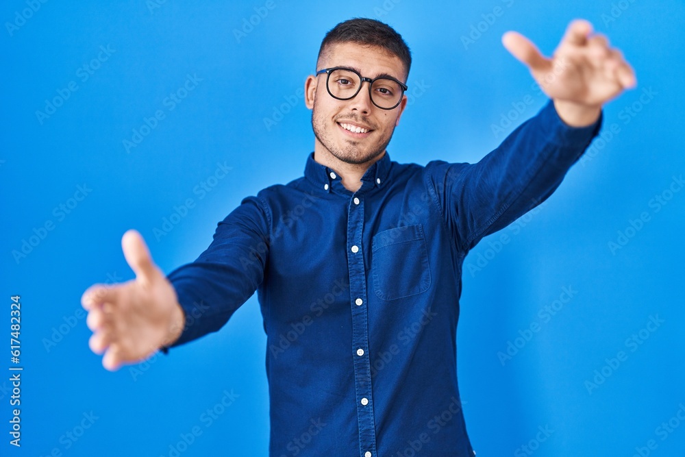Naklejka premium Young hispanic man wearing glasses over blue background looking at the camera smiling with open arms for hug. cheerful expression embracing happiness.