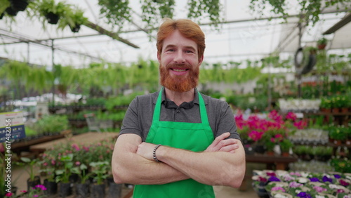 Young worker with arms crossed standing inside Flower Shop. A happy red hair male employee with green apron smiling at camera