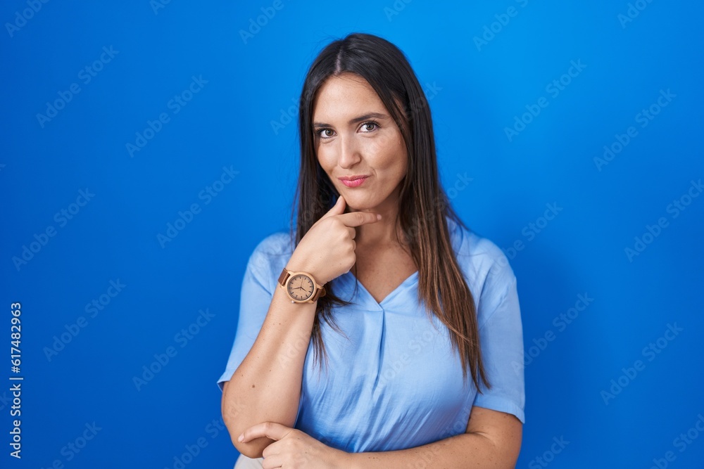 Young brunette woman standing over blue background looking confident at the camera smiling with crossed arms and hand raised on chin. thinking positive. Young brunette woman standing over blue background looking confident at the camera smiling with crossed arms and hand raised on chin. thinking positive.