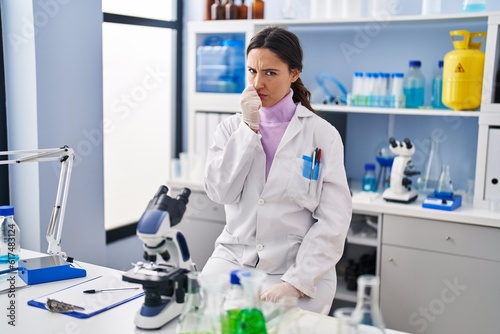 Young brunette woman working at scientist laboratory looking stressed and nervous with hands on mouth biting nails. anxiety problem.