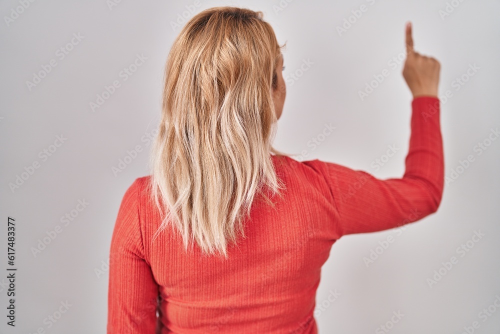 Blonde woman standing over isolated background posing backwards pointing ahead with finger hand Blonde woman standing over isolated background posing backwards pointing ahead with finger hand
