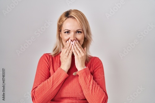 Blonde woman standing over isolated background laughing and embarrassed giggle covering mouth with hands, gossip and scandal concept