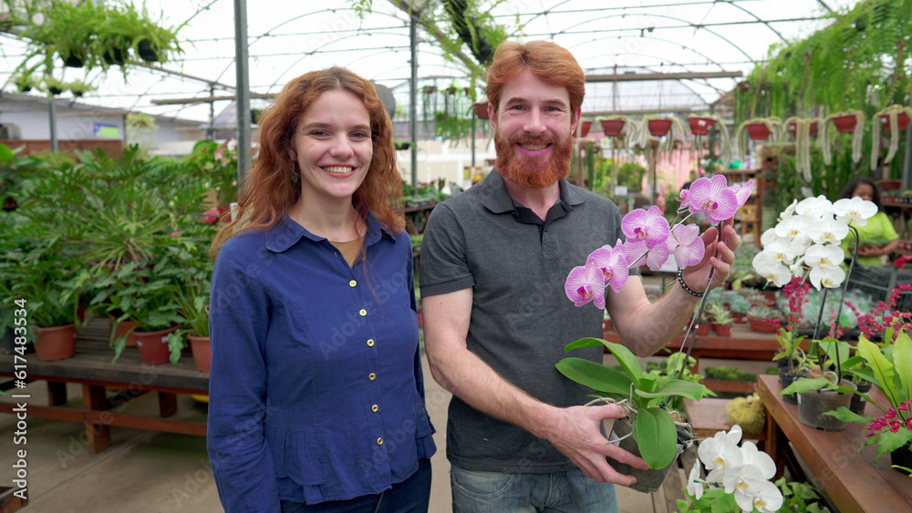Portrait of happy couple holding flower inside Plant Store while looking at camera smiling. A redhead man and woman shopping for decoration together