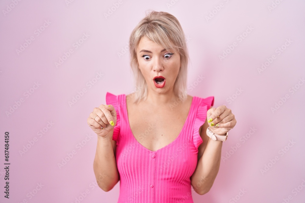 Young caucasian woman standing over pink background pointing down with fingers showing advertisement, surprised face and open mouth Young caucasian woman standing over pink background pointing down with fingers showing advertisement, surprised face and open mouth