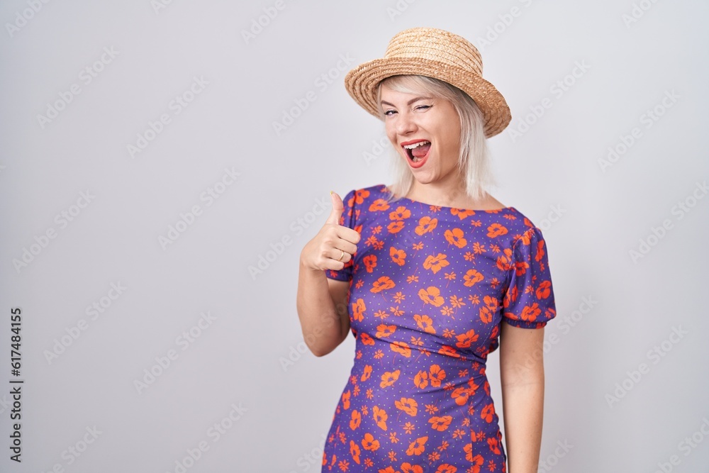Young caucasian woman wearing flowers dress and summer hat doing happy thumbs up gesture with hand. approving expression looking at the camera showing success. Young caucasian woman wearing flowers dress and summer hat doing happy thumbs up gesture with hand. approving expression looking at the camera showing success.