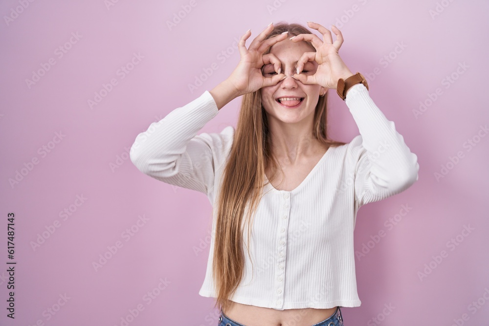 Young caucasian woman standing over pink background doing ok gesture like binoculars sticking tongue out, eyes looking through fingers. crazy expression. Young caucasian woman standing over pink background doing ok gesture like binoculars sticking tongue out, eyes looking through fingers. crazy expression.