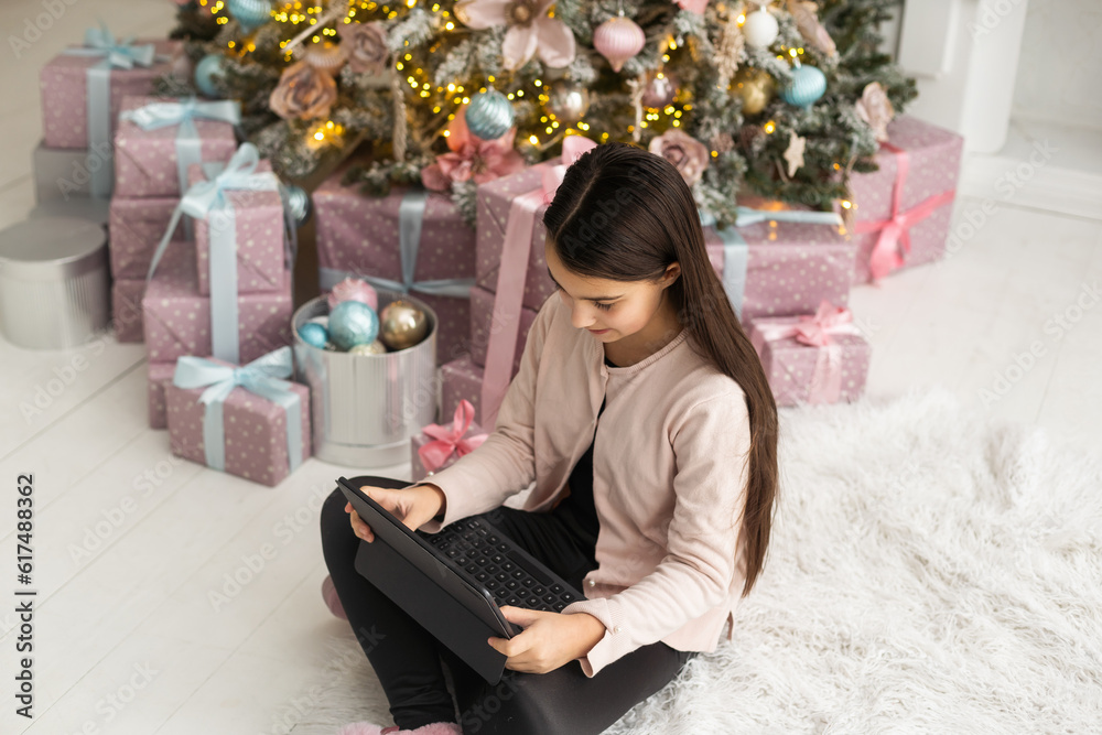 teenage girl works at home remotely with laptop on the background of a Christmas tree. Quarantine, work on holidays. atmosphere and work at home
