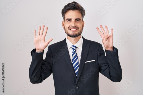 Young hispanic man with tattoos wearing business suit and tie showing and pointing up with fingers number nine while smiling confident and happy.