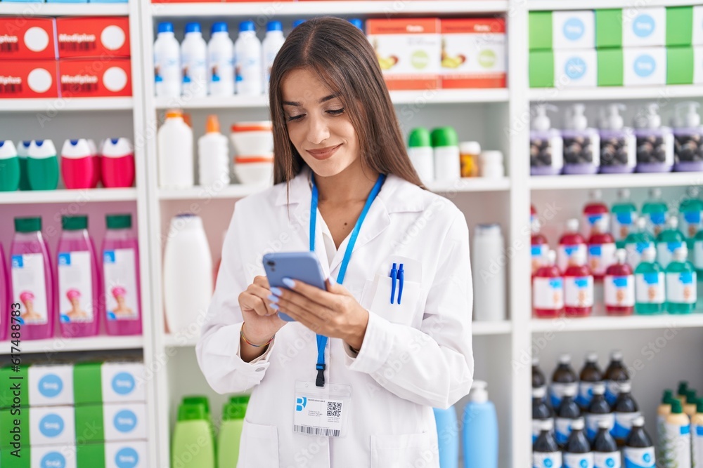 Young beautiful hispanic woman pharmacist using smartphone working at pharmacy