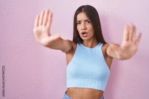 Young brunette woman standing over pink background doing stop gesture with hands palms  angry and frustration expression