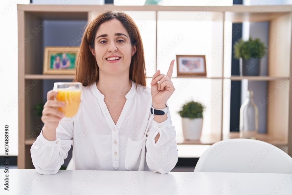 Brunette woman drinking glass of orange juice cheerful with a smile on face pointing with hand and finger up to the side with happy and natural expression Brunette woman drinking glass of orange juice cheerful with a smile on face pointing with hand and finger up to the side with happy and natural expression