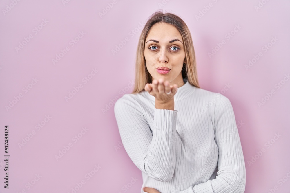 Young blonde woman wearing white sweater over pink background looking at the camera blowing a kiss with hand on air being lovely and sexy. love expression. Young blonde woman wearing white sweater over pink background looking at the camera blowing a kiss with hand on air being lovely and sexy. love expression.