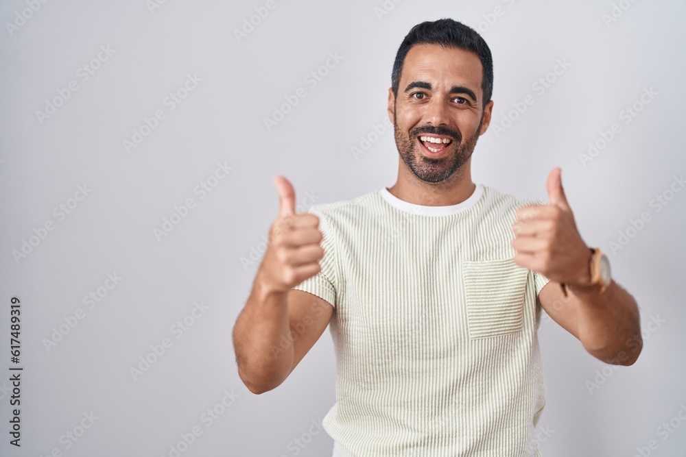 Hispanic man with beard standing over isolated background success sign doing positive gesture with hand, thumbs up smiling and happy. cheerful expression and winner gesture. Hispanic man with beard standing over isolated background success sign doing positive gesture with hand, thumbs up smiling and happy. cheerful expression and winner gesture.