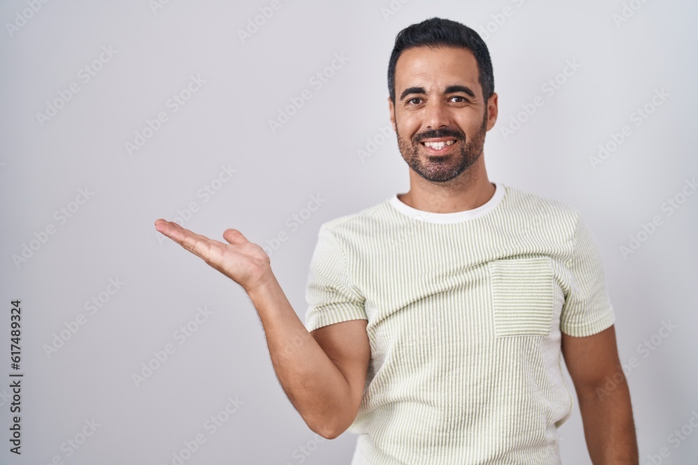 Hispanic man with beard standing over isolated background smiling cheerful presenting and pointing with palm of hand looking at the camera. Hispanic man with beard standing over isolated background smiling cheerful presenting and pointing with palm of hand looking at the camera.