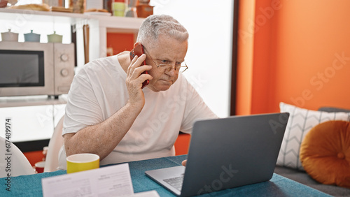 Middle age grey-haired man talking on smartphone using laptop at dinning room