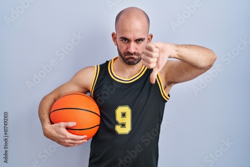 Young bald man with beard wearing basketball uniform holding ball looking unhappy and angry showing rejection and negative with thumbs down gesture. bad expression.