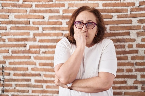 Senior woman with glasses standing over bricks wall looking stressed and nervous with hands on mouth biting nails. anxiety problem.