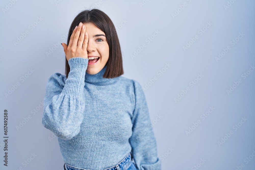 Young hispanic woman standing over blue background covering one eye with hand, confident smile on face and surprise emotion. Young hispanic woman standing over blue background covering one eye with hand, confident smile on face and surprise emotion.
