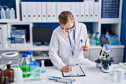Young blonde woman scientist writing report holding test tube at laboratory