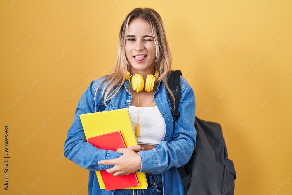 Young blonde woman wearing student backpack and holding books winking looking at the camera with sexy expression, cheerful and happy face.