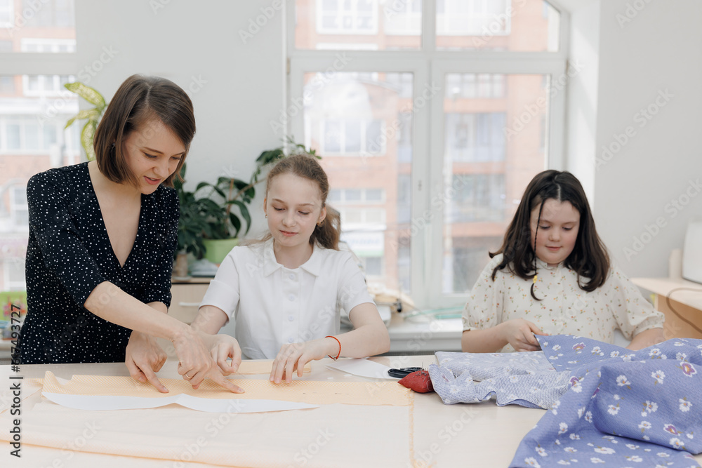 Dressmaker conducts sewing master class in school for two girl child. Smiling female designer or seamstress talking with children, education work Dressmaker conducts sewing master class in school for two girl child. Smiling female designer or seamstress talking with children, education work