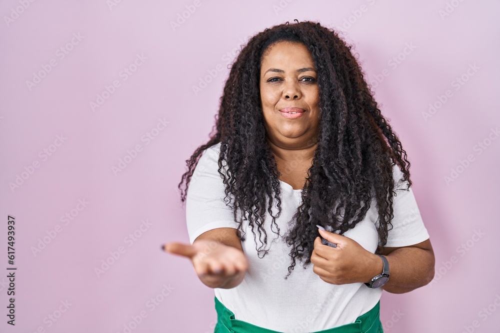 Plus size hispanic woman standing over pink background smiling cheerful offering palm hand giving assistance and acceptance. Plus size hispanic woman standing over pink background smiling cheerful offering palm hand giving assistance and acceptance.