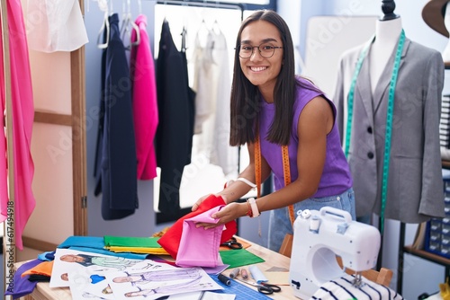 Young beautiful hispanic woman tailor smiling confident holding cloths at tailor shop
