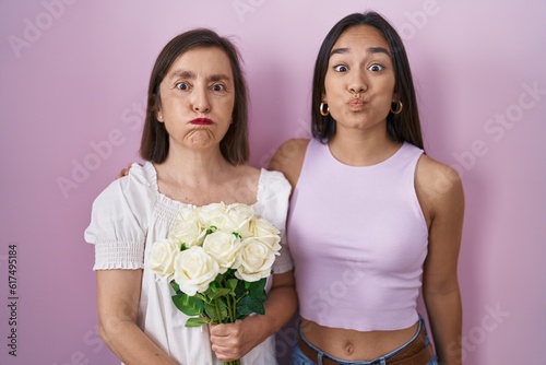 Hispanic mother and daughter holding bouquet of white flowers puffing cheeks with funny face. mouth inflated with air, crazy expression.