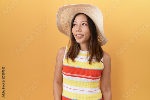 Middle age chinese woman wearing summer hat over yellow background looking away to side with smile on face, natural expression. laughing confident.
