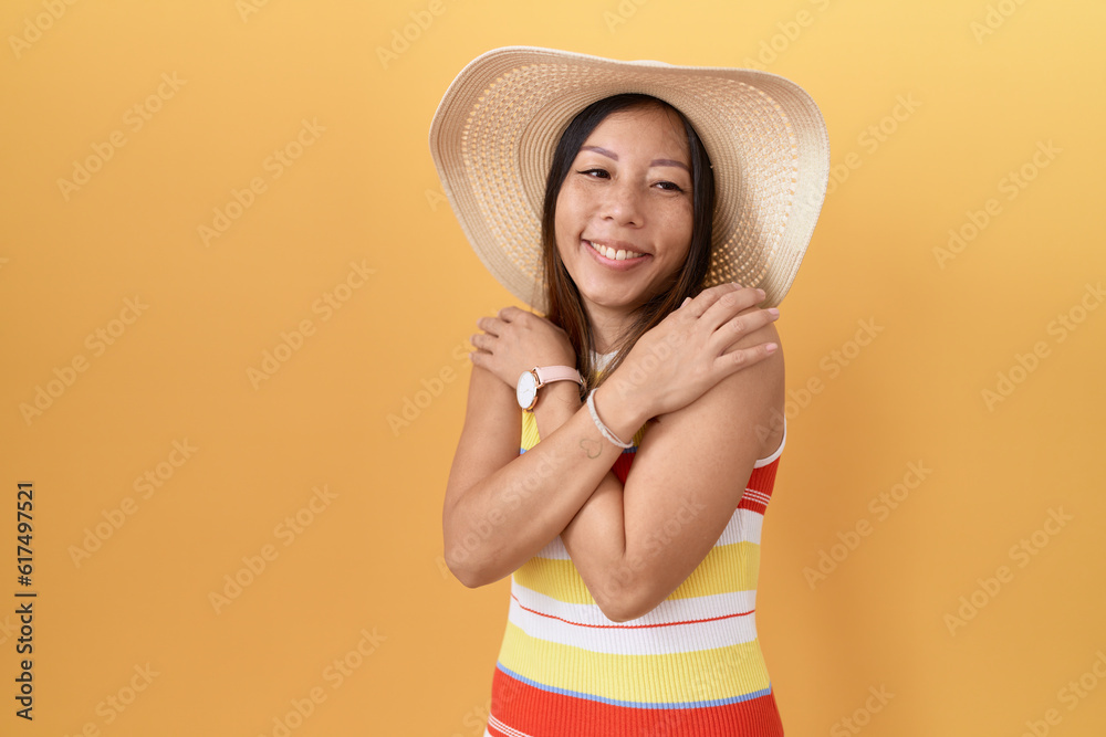 Middle age chinese woman wearing summer hat over yellow background hugging oneself happy and positive, smiling confident. self love and self care Middle age chinese woman wearing summer hat over yellow background hugging oneself happy and positive, smiling confident. self love and self care