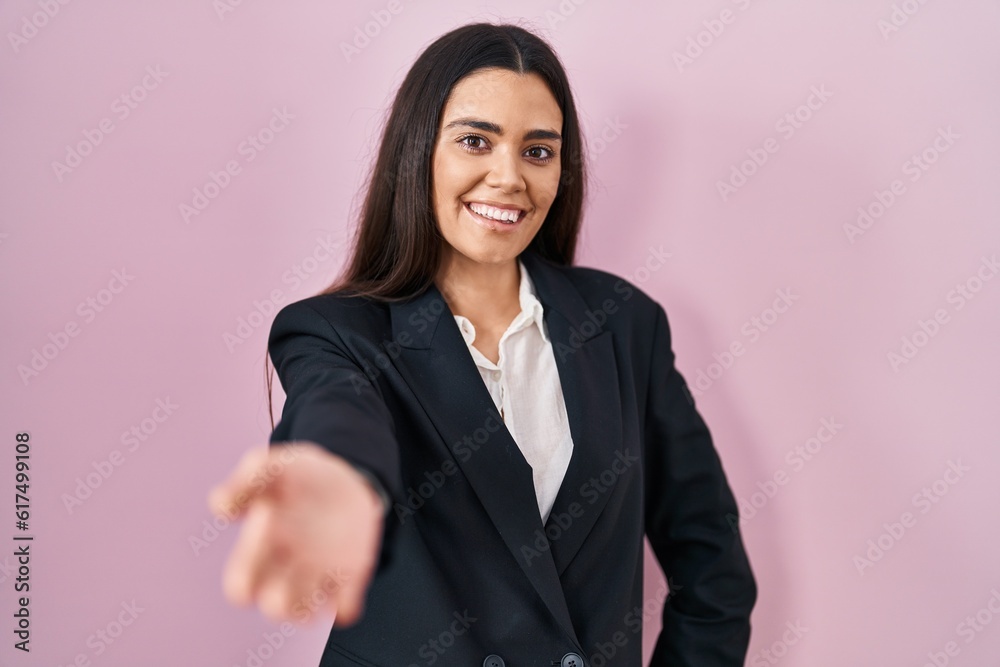 Young brunette woman wearing business style over pink background smiling friendly offering handshake as greeting and welcoming. successful business. Young brunette woman wearing business style over pink background smiling friendly offering handshake as greeting and welcoming. successful business.