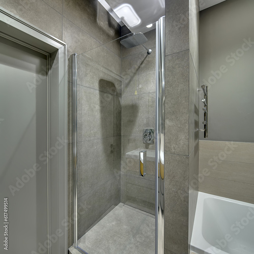 Contemporary bathroom with long tiles on the walls and floor. There is a shower cabin with a glass door  white bath  door  chrome towel holder  glowing lamps. Vertical.