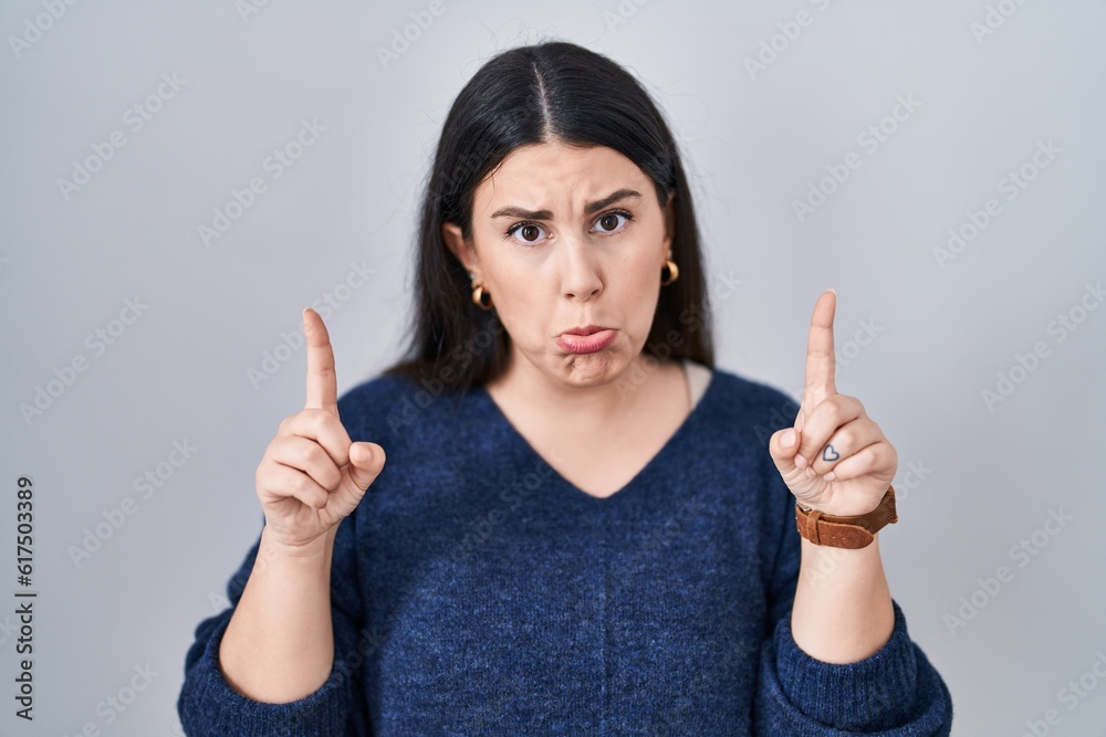Young brunette woman standing over isolated background pointing up looking sad and upset, indicating direction with fingers, unhappy and depressed. Young brunette woman standing over isolated background pointing up looking sad and upset, indicating direction with fingers, unhappy and depressed.