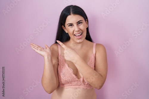 Young hispanic woman wearing pink bra amazed and smiling to the camera while presenting with hand and pointing with finger.